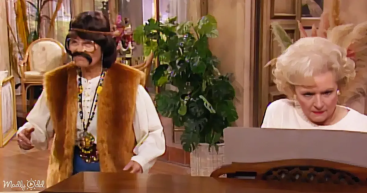 A man in a fur vest and wig stands beside a seated woman in a white outfit, both in a room with plants and furniture.