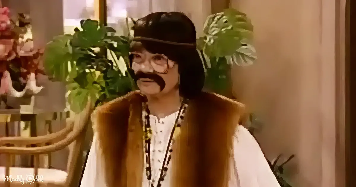 A person in a brown vest, white shirt, and black hat with a mustache stands in a room with plants and flowers.