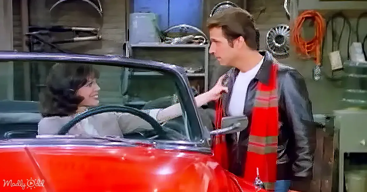 A man in a garage talks to a woman in a red car, surrounded by tools and equipment.
