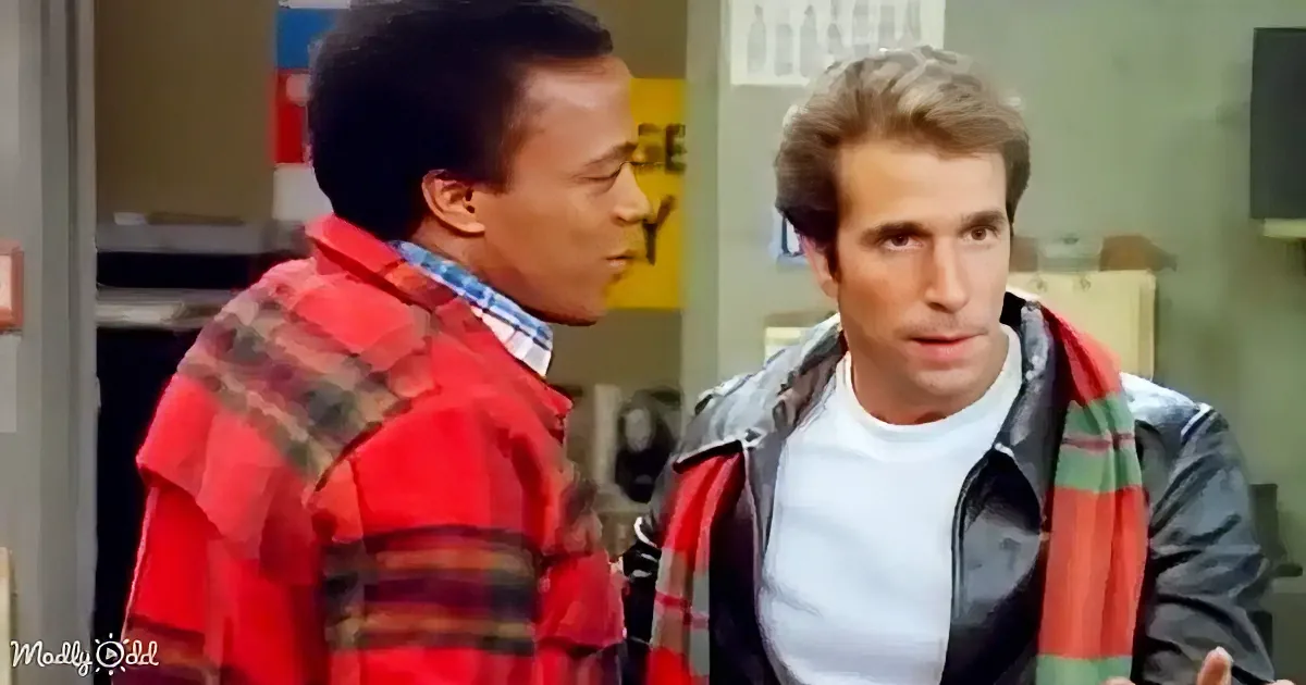A man in a red plaid shirt is talking to another man in a white t-shirt and black jacket in a room.
