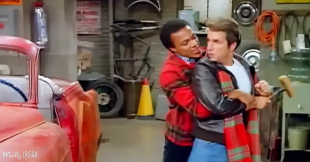 Two men in a garage, one in a red jacket holding the other, with a red car and various tools in the background.