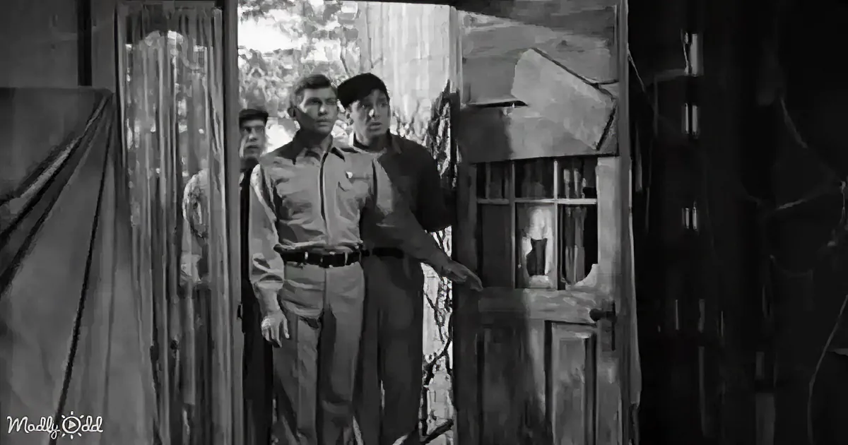 Two men in uniform stand by a wooden door, one gesturing, in a dimly lit setting.