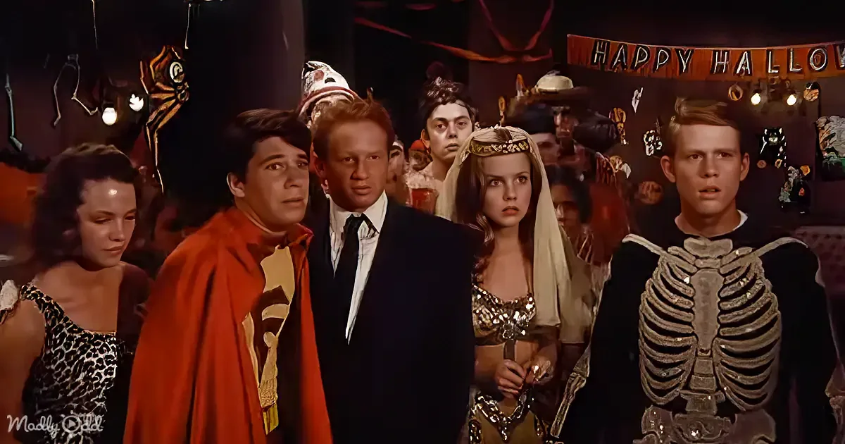 A group of people in costumes, including a skeleton, at a Halloween party with a "Happy Halloween" banner.