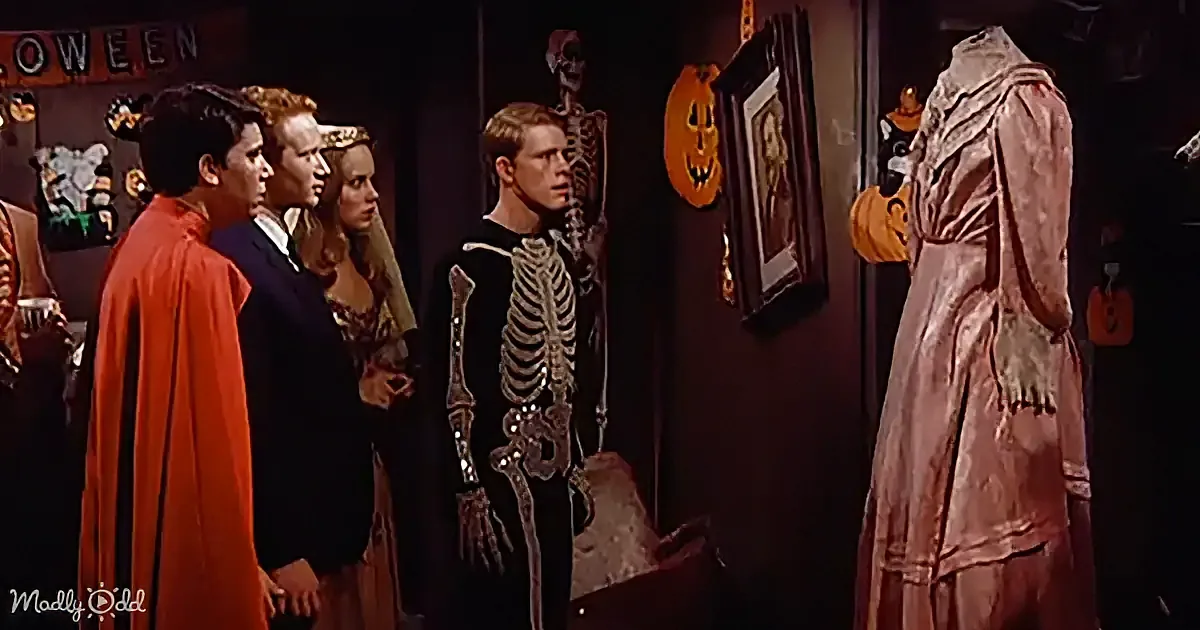 A man in a skeleton costume stands in front of a group of people in Halloween costumes, with decorations in the background.