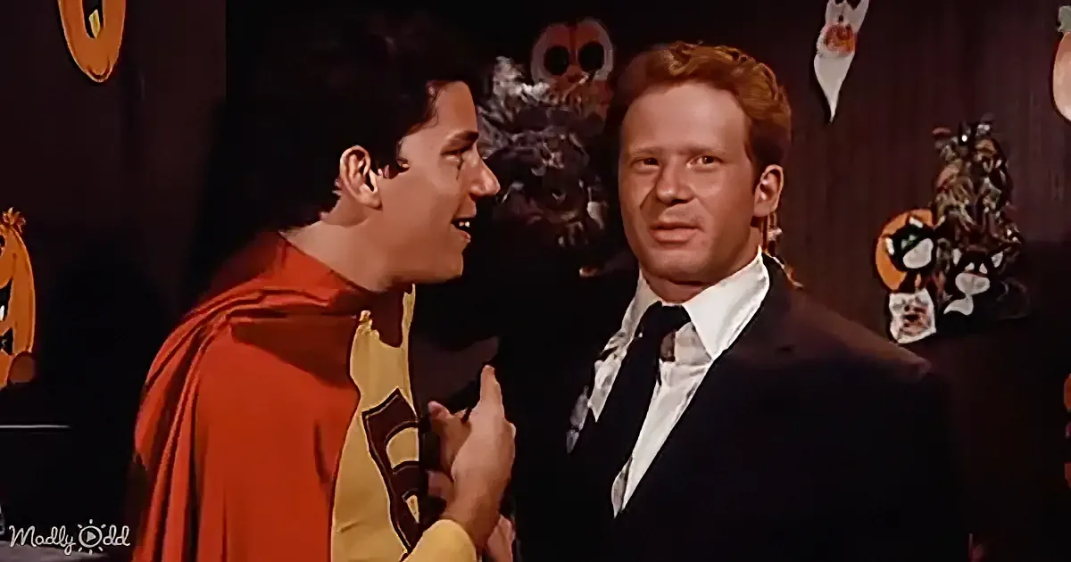 A man in a superhero costume is playfully interacting with another man in a suit, both smiling.