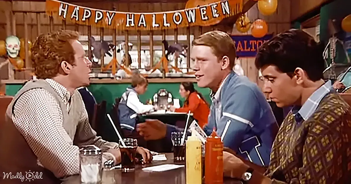 Three men sit at a table in a Halloween-decorated diner, engaged in conversation.