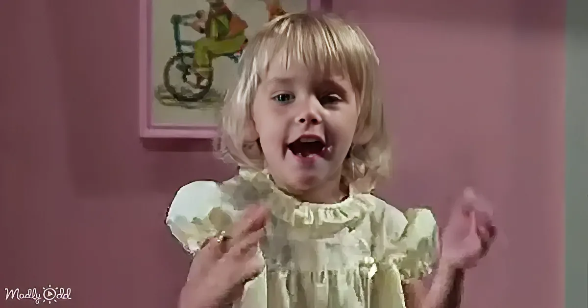 A young child with blonde hair and a green dress stands in front of a pink wall, gesturing with their hands.