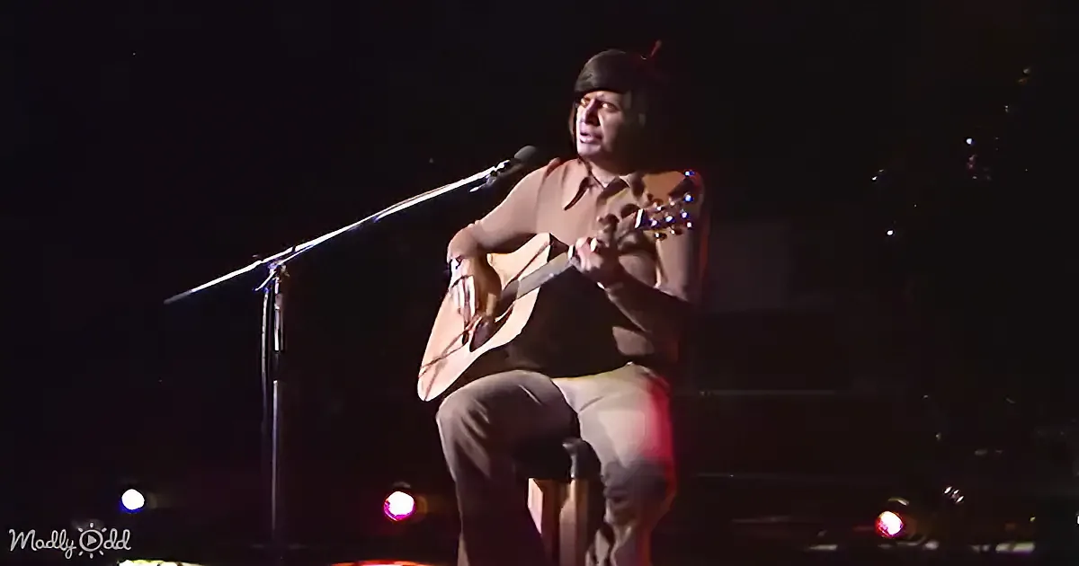 A man sits on a stool, playing an acoustic guitar and singing into a microphone.