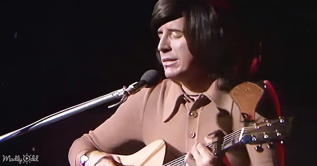 A person with shoulder-length hair plays an acoustic guitar and sings into a microphone.