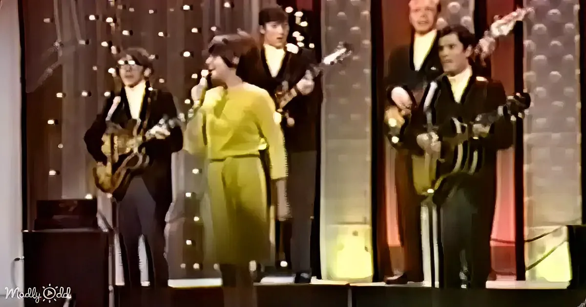 A group of performers, including a woman in a yellow dress, sing and play instruments on a stage with a backdrop of lights.