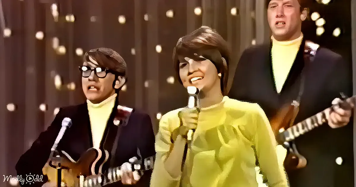 A person in a yellow outfit sings into a microphone, flanked by two guitarists in a retro setting.