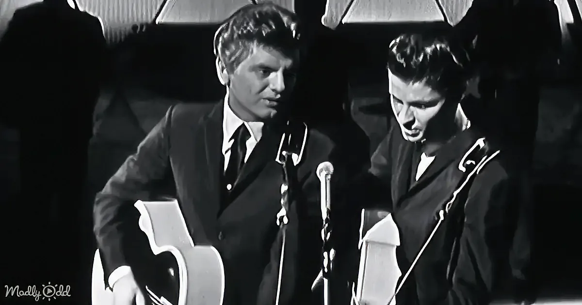 Two men in suits play guitars on stage, one with a microphone, in a black-and-white image.
