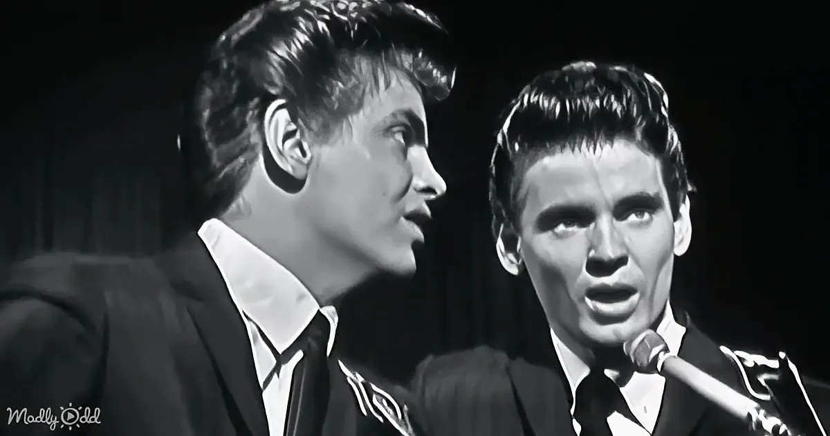 Two men in suits sing into a microphone, one with a microphone in front of him.