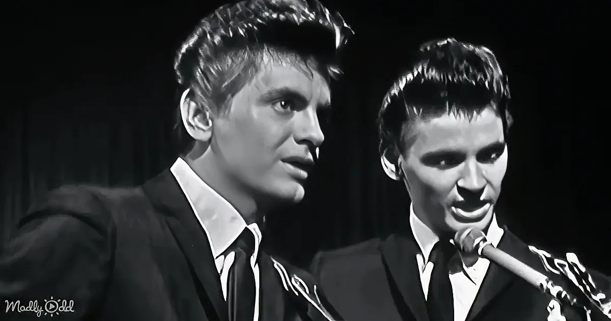 Two men in suits sing into microphones, one with a serious expression, the other with a smile.
