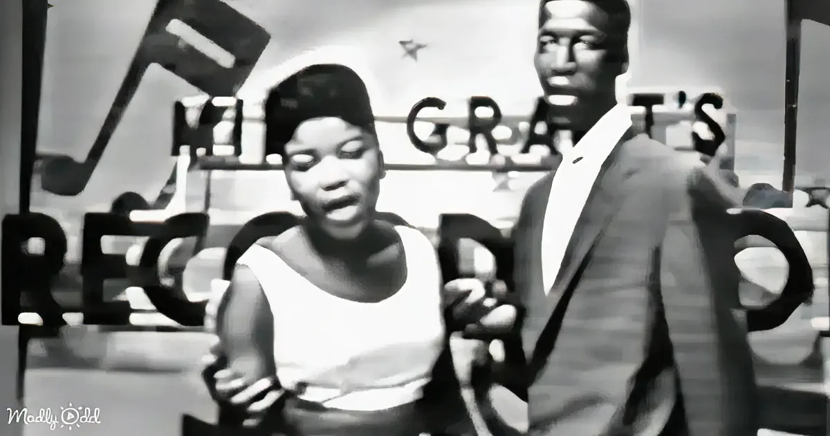 A woman in a white dress holds a microphone, standing in front of a sign that reads "M.GRAT'S RECORDS."