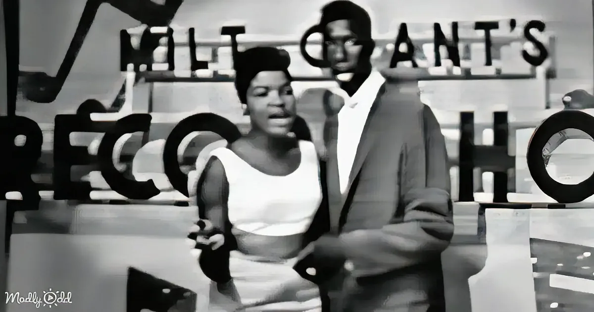 A woman in a white dress and a man in a suit stand in front of a sign that reads "Milt Grant's."