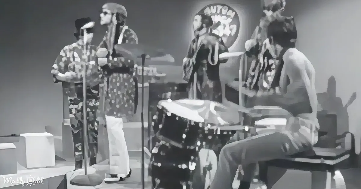 A band in 1960s attire performs on stage with microphones, drums, and a circular backdrop.