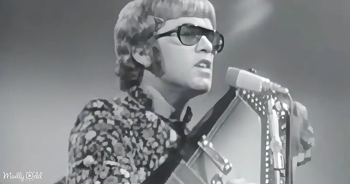 A person in a floral shirt and sunglasses plays a guitar on stage, singing into a microphone.