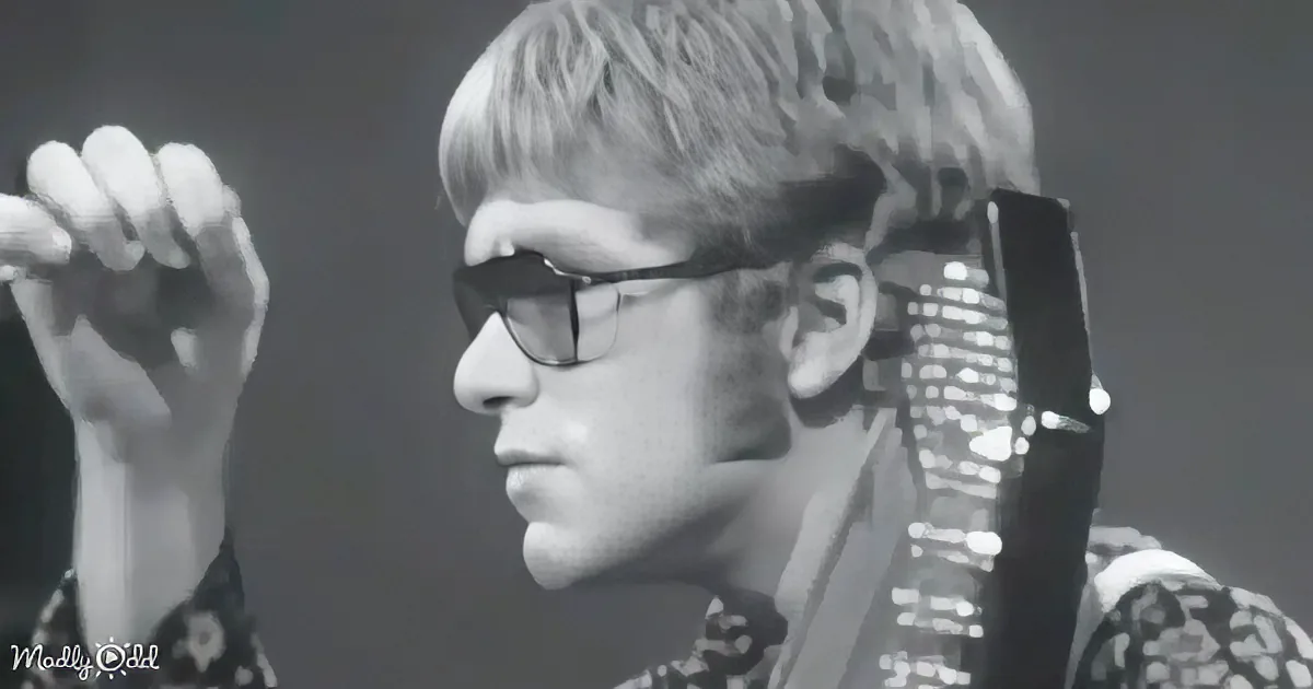 A person wearing glasses and a patterned shirt holds a guitar, with a hand raised near their face.