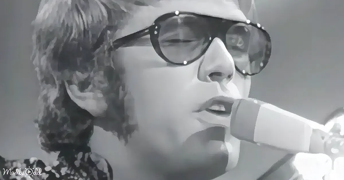 A person wearing sunglasses and a hat sings into a microphone, with a black-and-white filter.