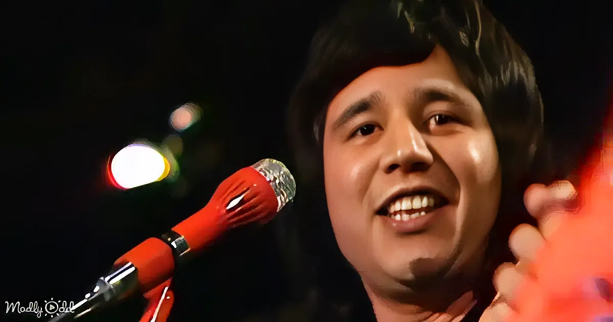A person with dark hair smiles near a red microphone, with blurred lights in the background.