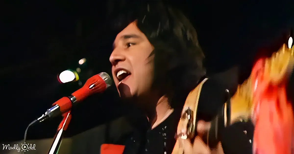 A person with long hair sings into a microphone while holding a guitar, with colorful stage lights in the background.
