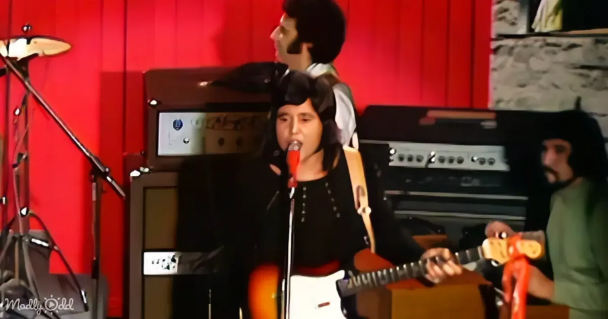 A person plays guitar and sings into a microphone in front of a red wall with musical equipment.