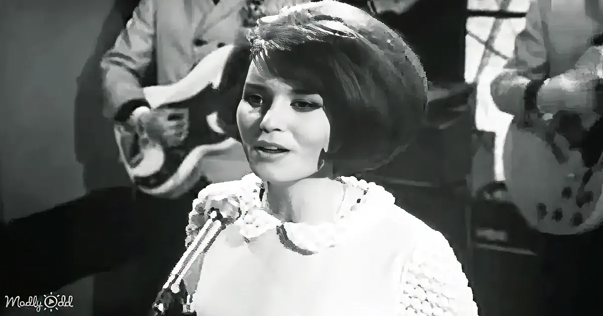 A woman in a textured white outfit sings into a microphone, surrounded by musicians.