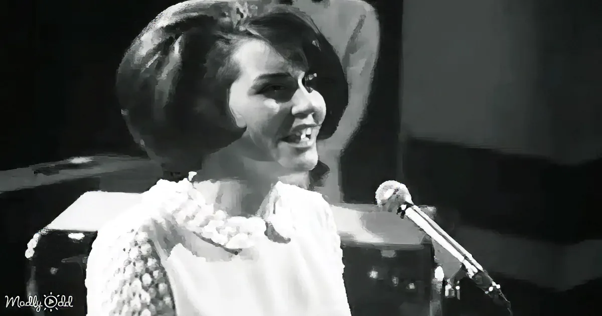 A woman in a white dress with a floral pattern sings into a microphone, smiling.