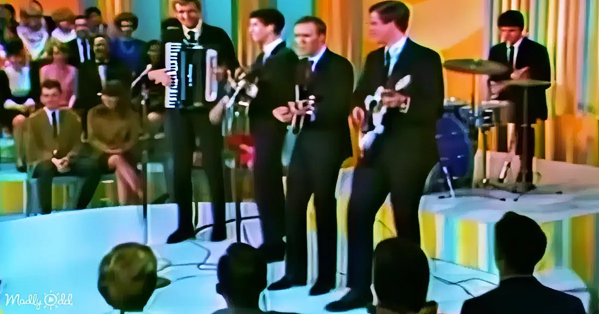 A group of men in suits perform on stage with microphones and instruments, surrounded by an audience and colorful backdrop.