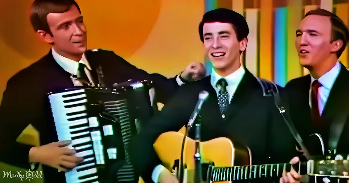A man plays an accordion while two others sing and play guitar in a vibrant, colorful setting.