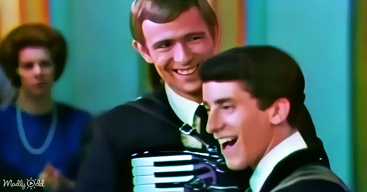 A man in a suit sings into a microphone, smiling, with another person behind him.