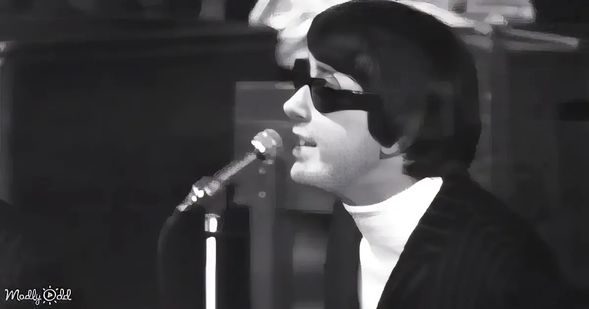 A person wearing glasses and a suit sings into a microphone on stage.