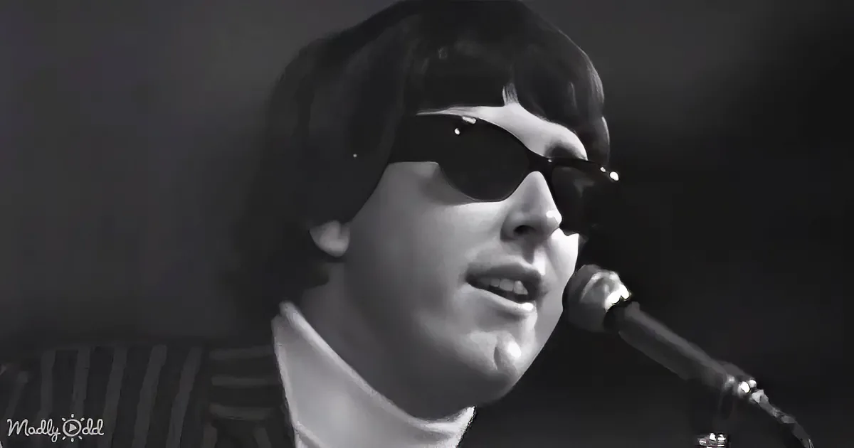 A person wearing sunglasses and a high-collared shirt sings into a microphone, set against a dark background.