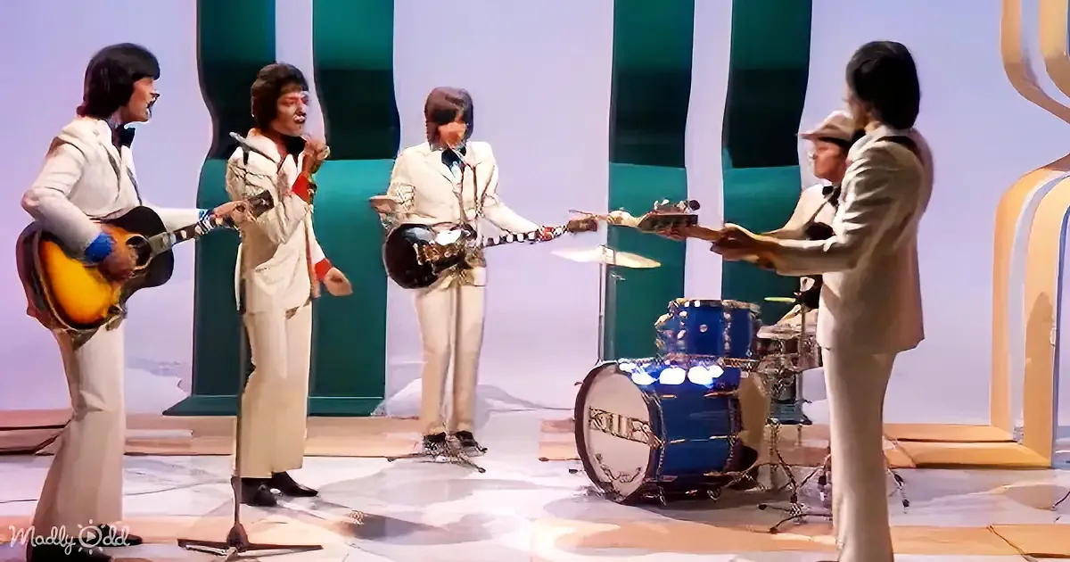 A band in white suits performs on stage, with a drummer and guitarists singing and playing.