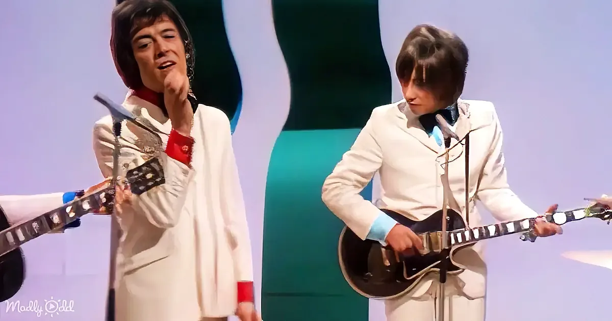 A man in a white suit plays a guitar on stage, another man sings nearby.