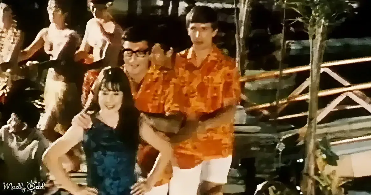 A man in an orange shirt carries a child, surrounded by people in a lively outdoor setting.