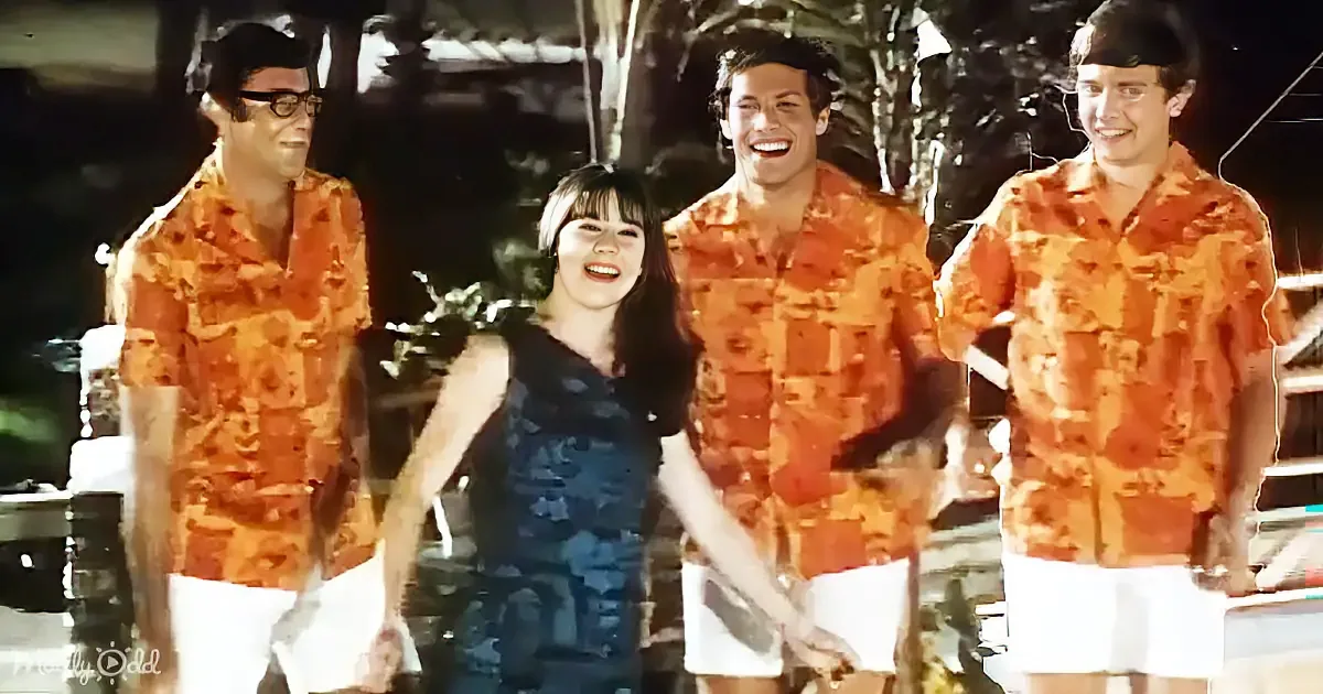 A woman in a blue dress stands with three men in matching orange shirts, smiling in a tropical setting.