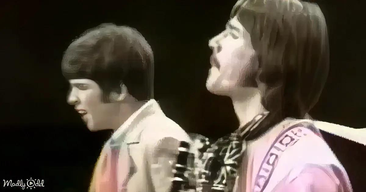 A man with long hair and a guitar sings passionately on stage, accompanied by another musician.