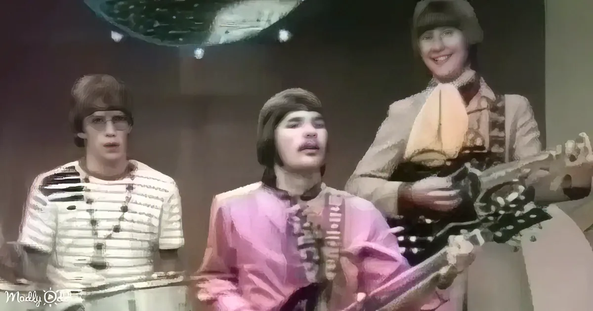 A man in a pink suit plays guitar while two others sing and play instruments in a vintage setting.