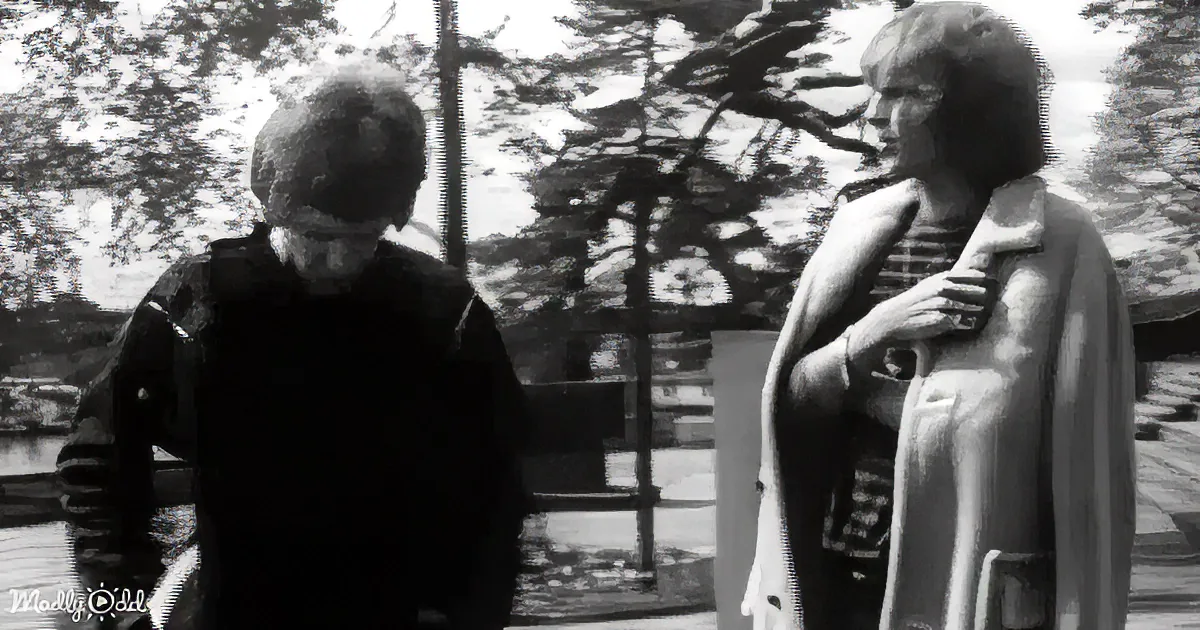 A person in a dark coat walks past another person in a light coat holding a baby, with trees and a fence in the background.
