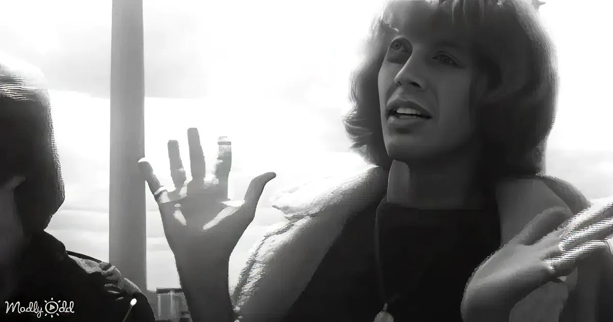 A woman with short hair gestures while speaking, wearing a dark jacket, in a black-and-white image.
