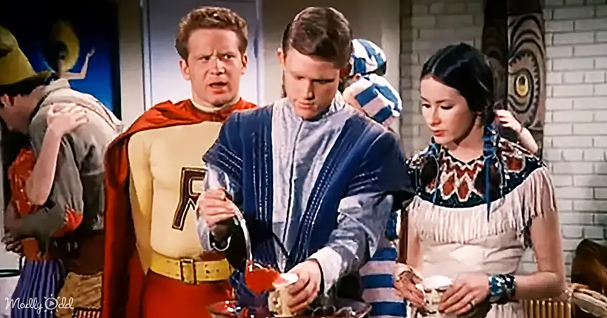 A man in a blue robe pours a drink while a woman in a Native American-style dress looks on, with another man in a...