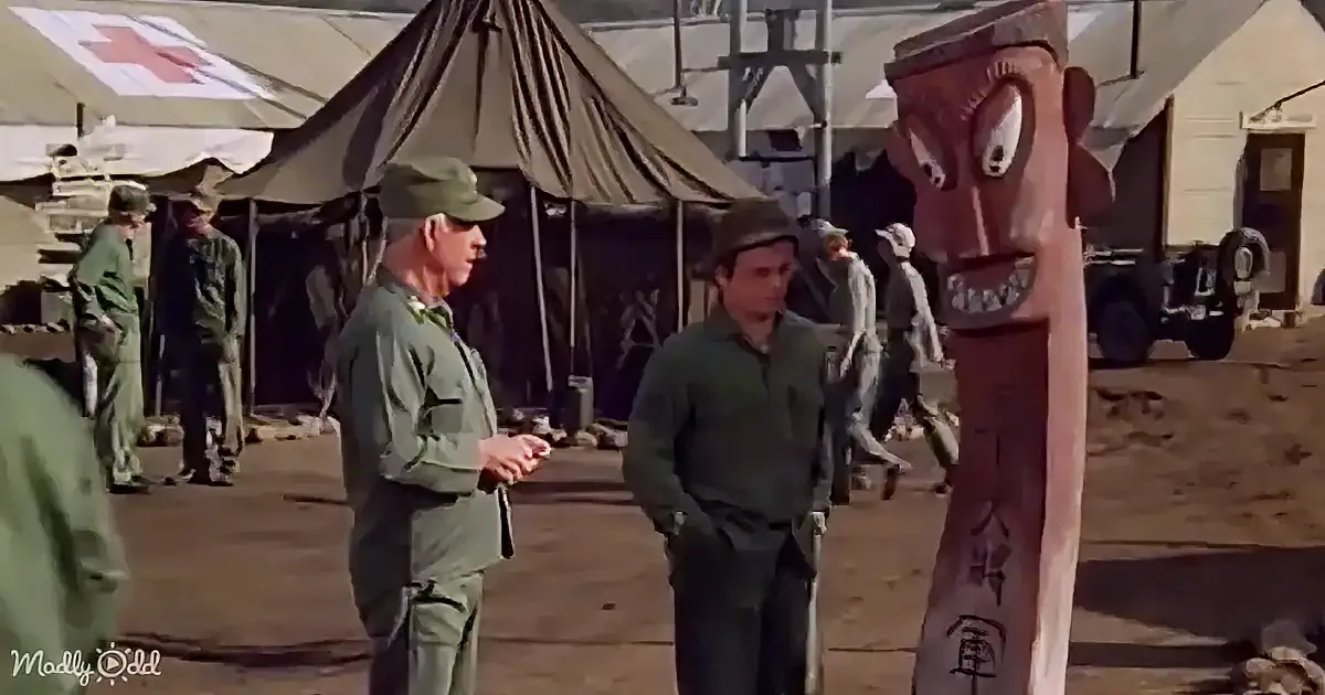 Two men in military uniforms converse in a camp setting with tents and vehicles in the background.