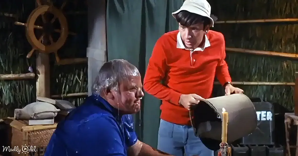 A man in a red sweater pours water on another man in a blue shirt, both in a rustic setting.