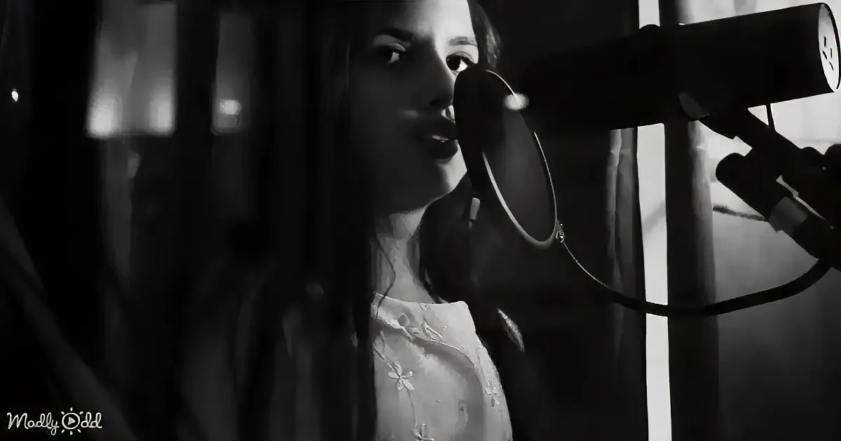 A woman sings into a microphone in a dimly lit room, wearing a light-colored top.