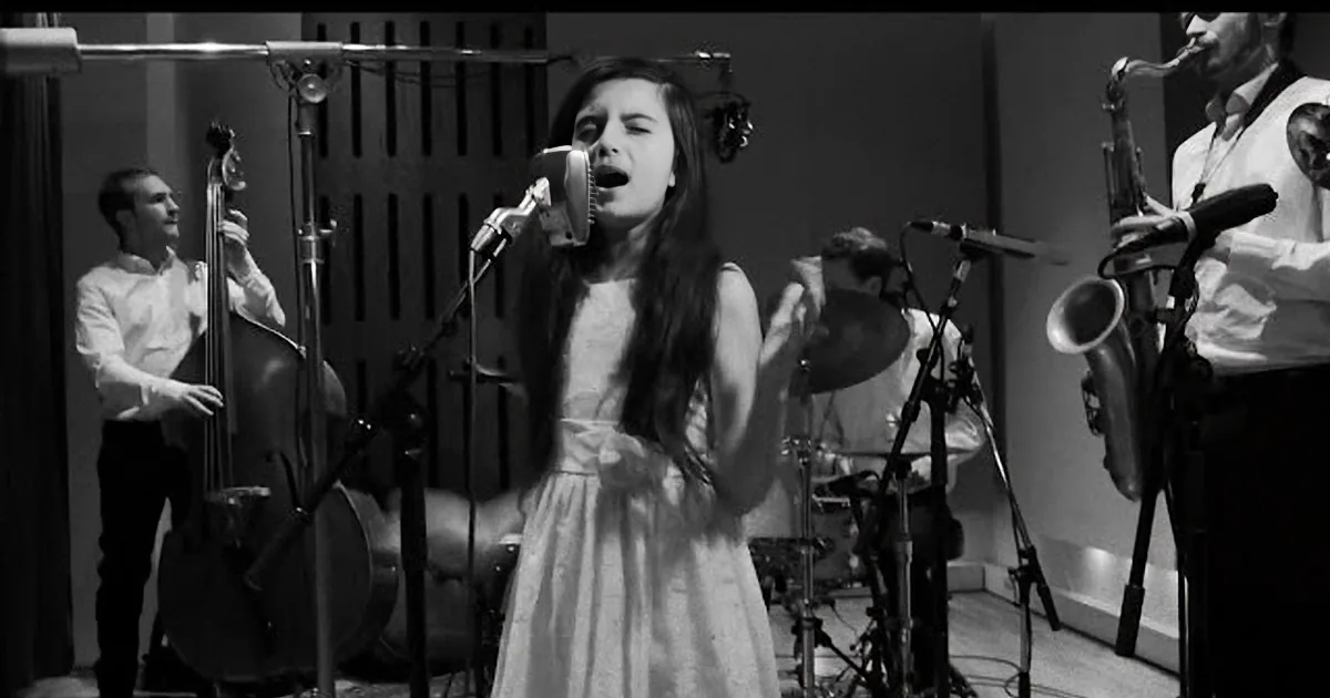 A young woman in a white dress sings into a microphone, surrounded by musicians in a recording studio.