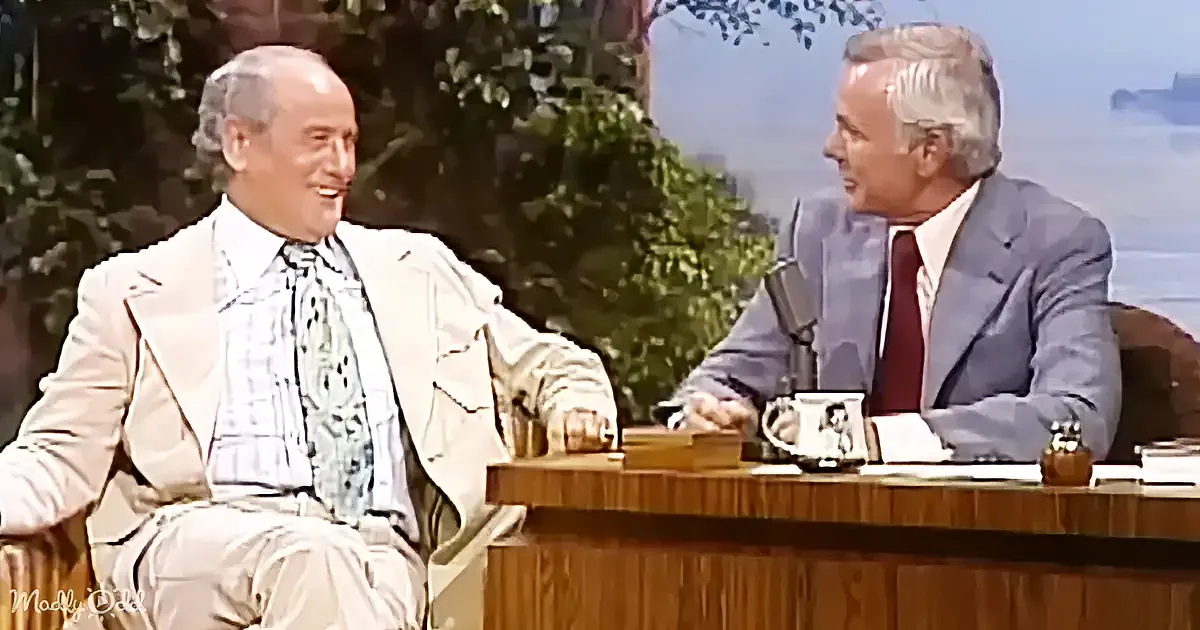 A man in a beige suit and patterned tie sits on a couch, smiling at another man in a blue suit seated at a desk.