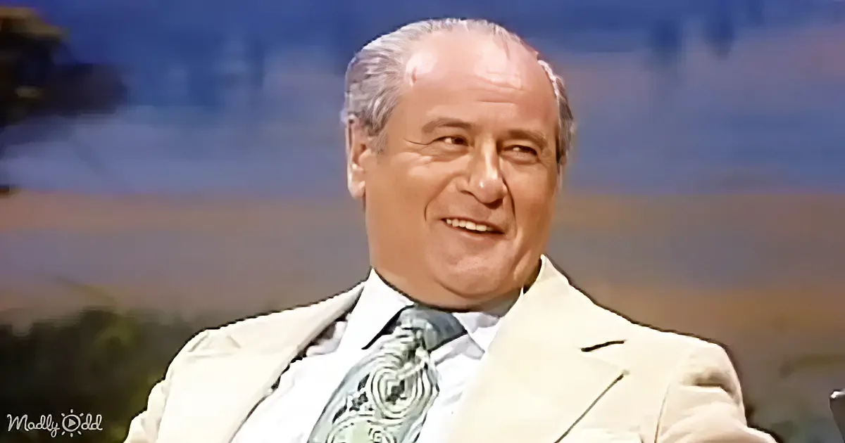 A man in a light suit and patterned tie is smiling, seated against a blurred background.