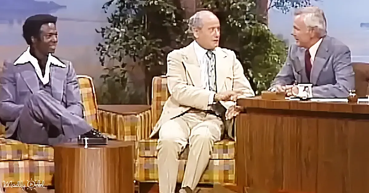 A man in a light suit sits on a plaid chair, engaged in conversation with two others on a talk show set.